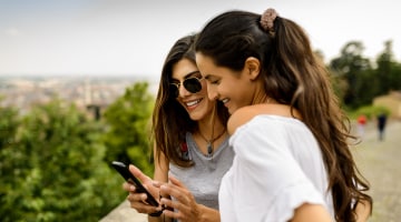 Two young South Asian women in short-sleeved shirts who are standing outdoors smile and look down at a cell phone.