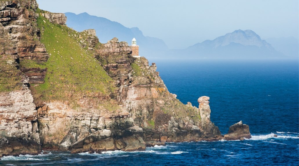Cape Point is made up of craggy light-brown cliffs. Grass shades one face and a red brick lighthouse perches over the deepest decline.