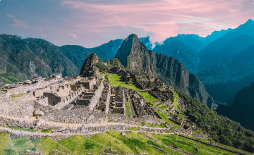 The historic sanctuary of Machu Picchu curves along the top of a grassy mountain, with more peaks rising into pink clouds beyond.