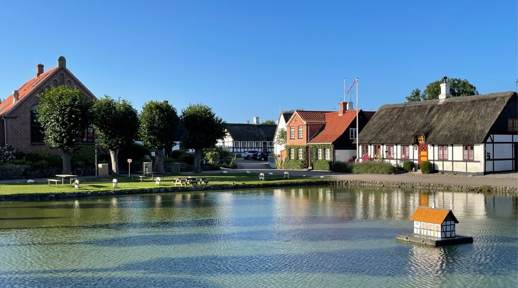 A duck house sits in the middle of a pond. Nearby are half-timbered houses and a park set with picnic tables and benches.