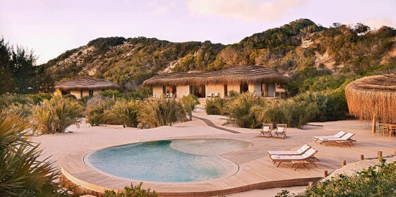 A private residence with an overhanging thatched roof blends almost seamlessly into a nearby hill. A private pool is flanked by sun loungers.