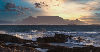 Waves crash on a rocky shoreline. Just across the water, a flat-topped mountain is dramatically silhouetted against a sunset.