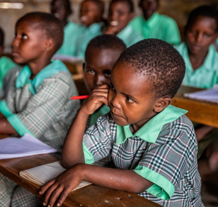 In a primary school, rows of young boys with short-cropped hair sit at benches with pencils and paper, all dressed in green uniforms.