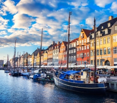 On a sunny day, sailboats are docked next to a row of brightly painted buildings with umbrella-shaded seating out front.