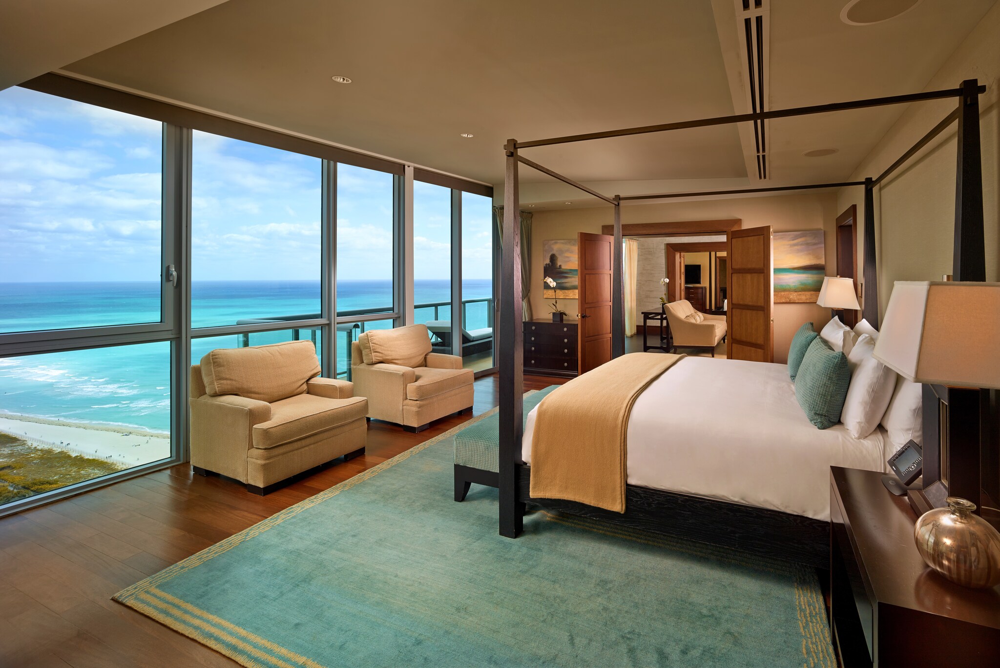 In a suite, a big bed faces a row of floor-to-ceiling windows. Doors on one end of the room open into a living space.