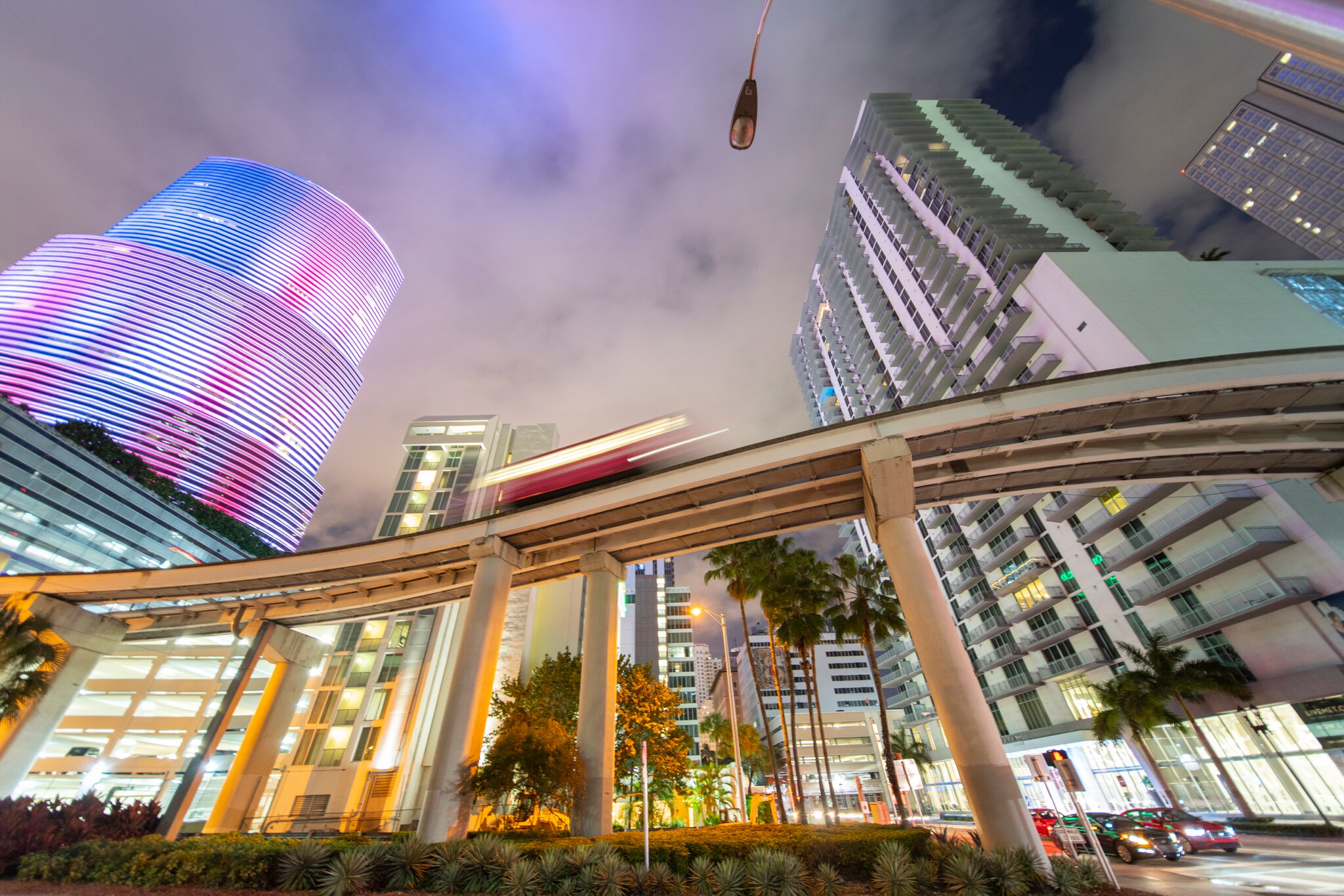 Looking up from below at night, an elevated light rail tram speeds pas the Brickell City Centre, which is lit up in bright blue and pink.
