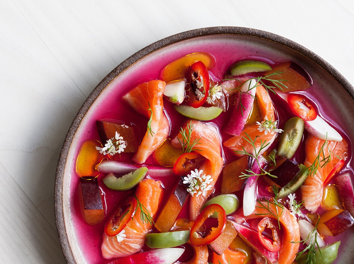 Close-up overhead view of a dish of thinly-slided fish, olives, fruit,and herbs, floating in a fuchsia liquid.