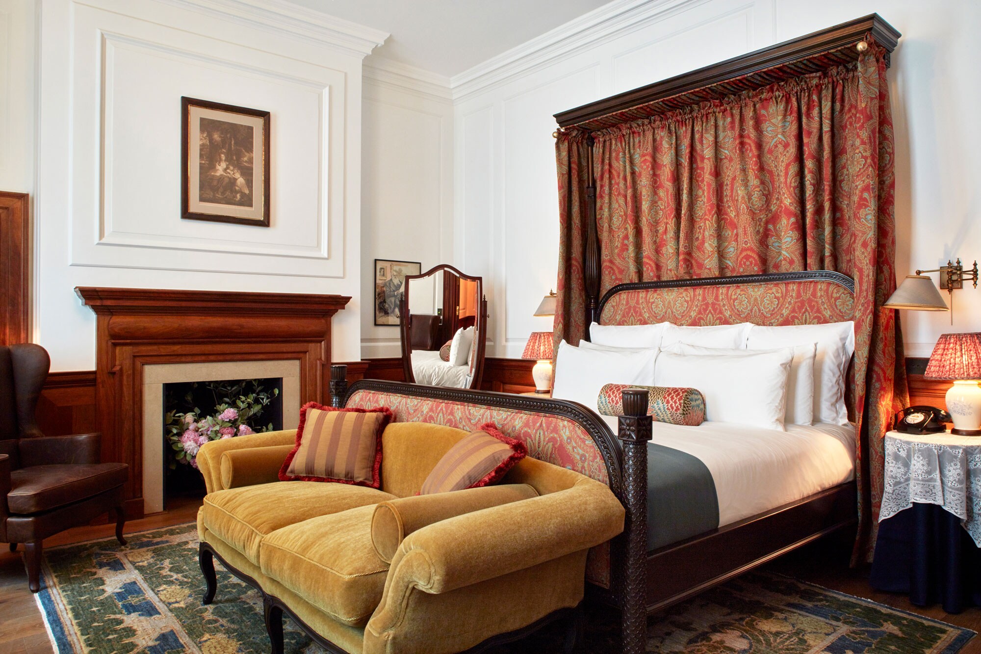 A hotel room features a large bed with red-and-gold head- and foot-boards. A mustard couch is at its foot, with a fireplace to the side.