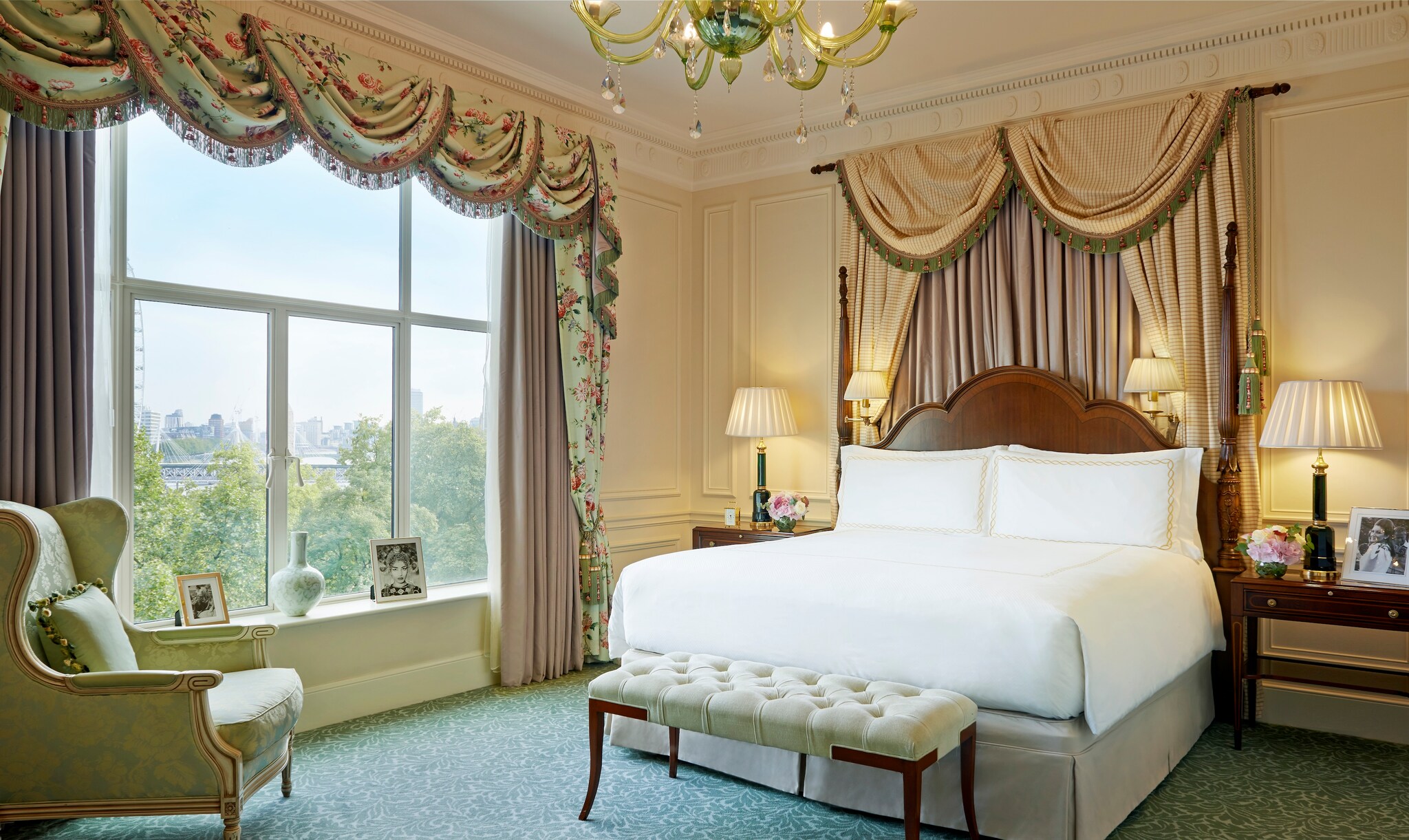A hotel room features a large bed under beige and cream curtains, brightly lit from wide windows along one wall.