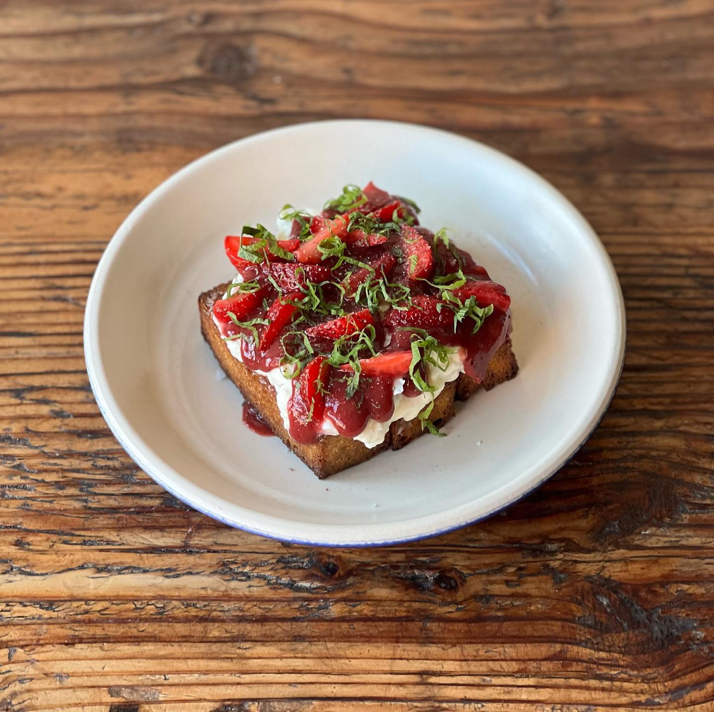 On a small round dish, a square pastry is covered in burrata, strawberries, red sauce, and curls of shredded herbs.