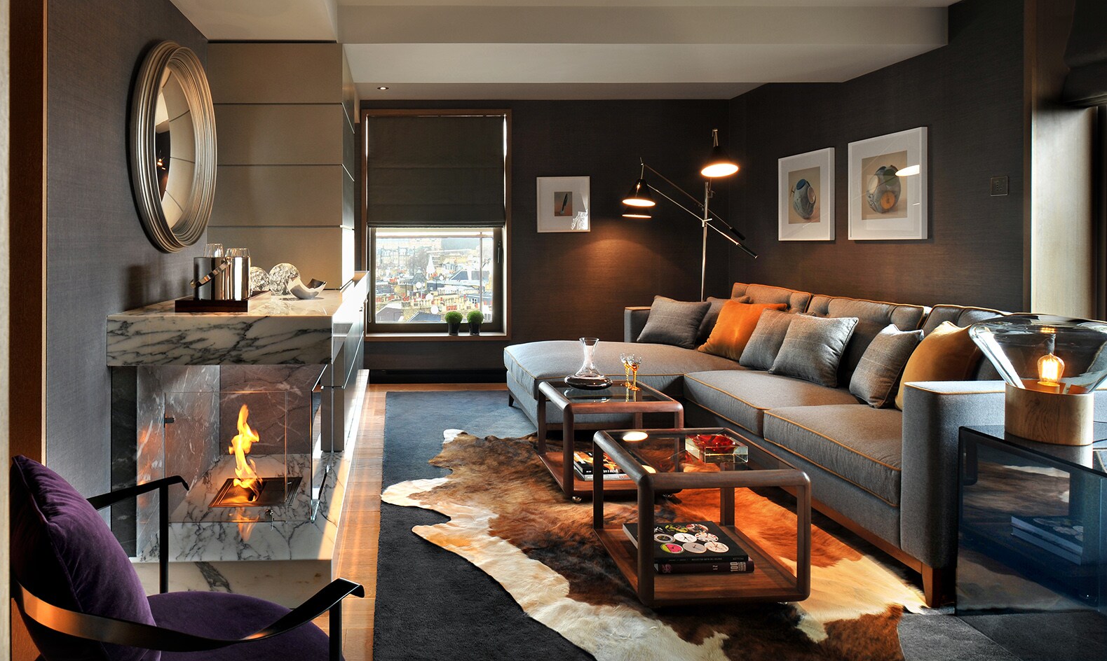 The living room of a suite features a long gray couch in front of a glassed-off fireplace. An animal skin rug lies under two coffee tables.
