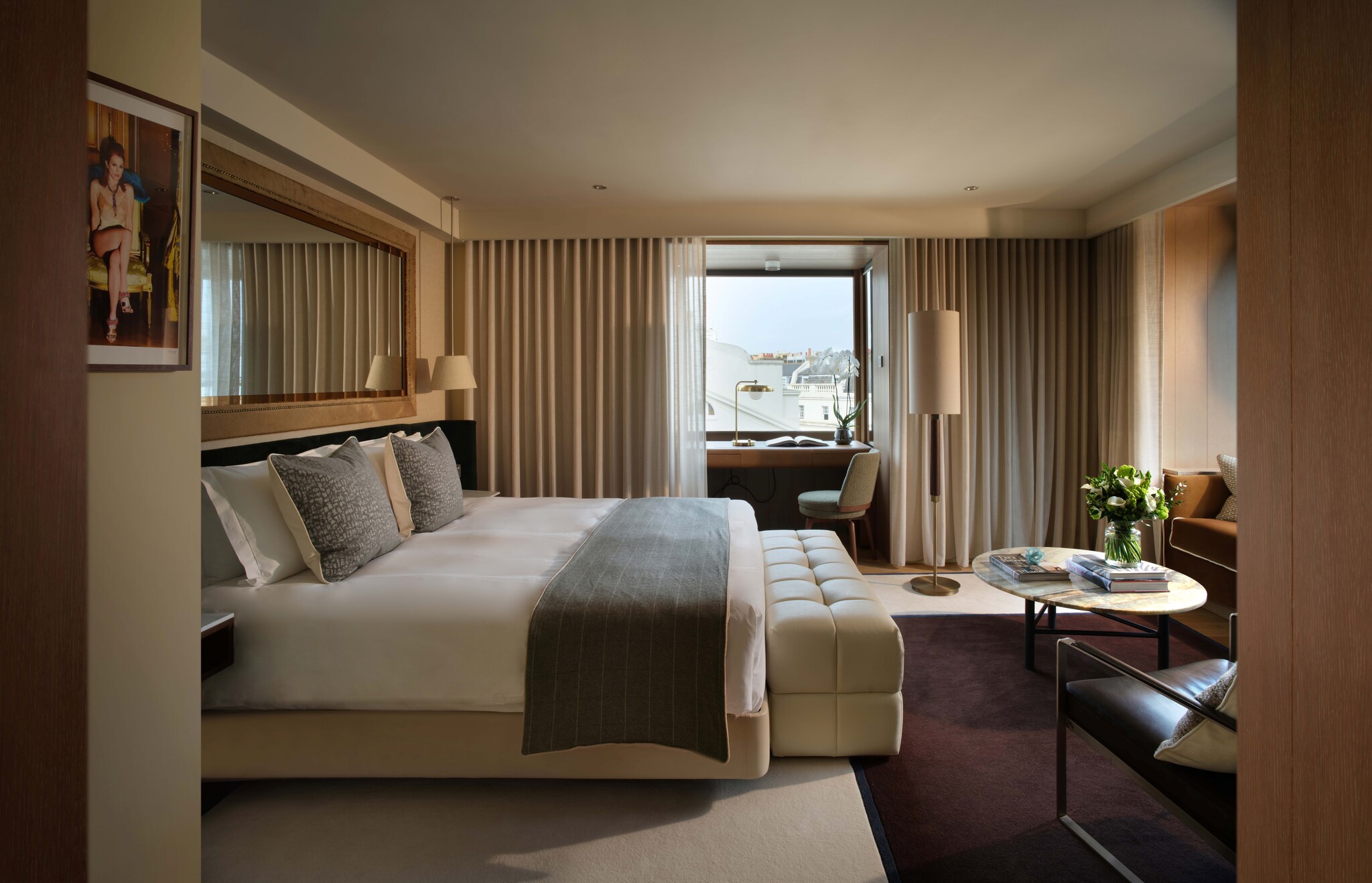 In a hotel room, a king-sized bed faces a window seat. Beyond it, past some curtains, a small desk sits in a nook below a window.