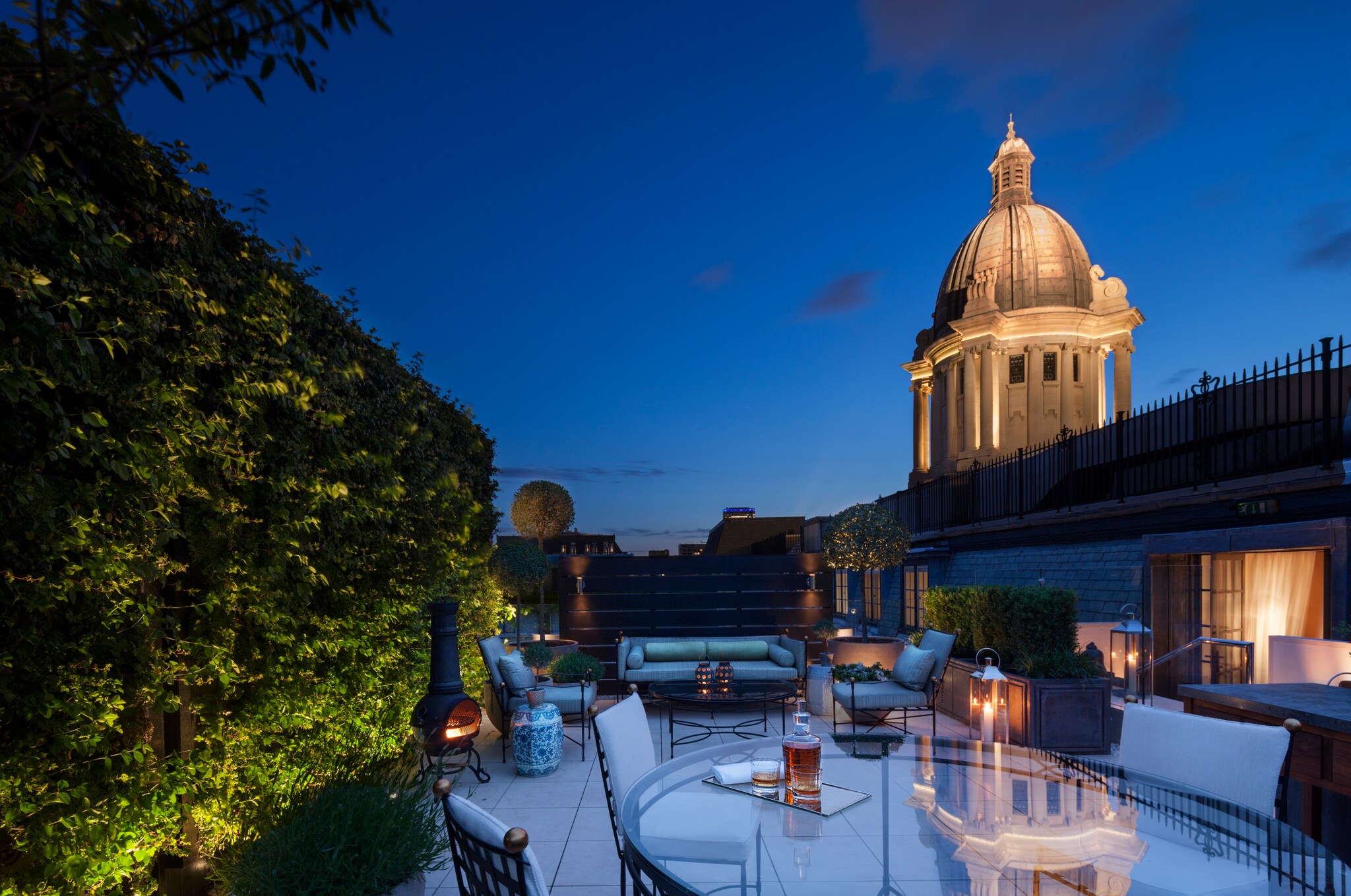 The Garden House is covered in greenery and adjoins a terrace with seating and a fireplace, the lit rotunda visible behind in the night.