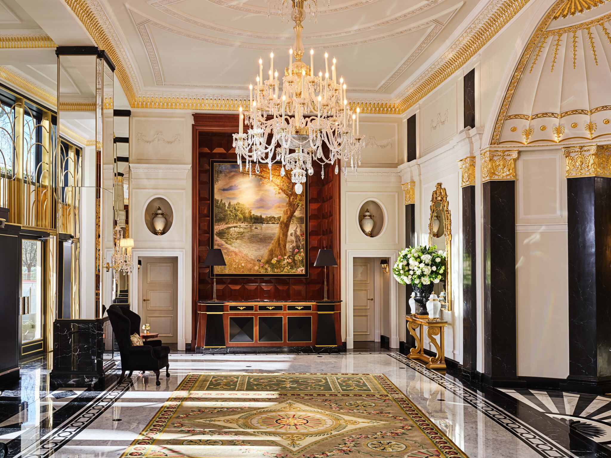 The hotel lobby features  mosaic floors, gilded crown molding, an ornate chandelier, and black marble finishes on pillars and walls.