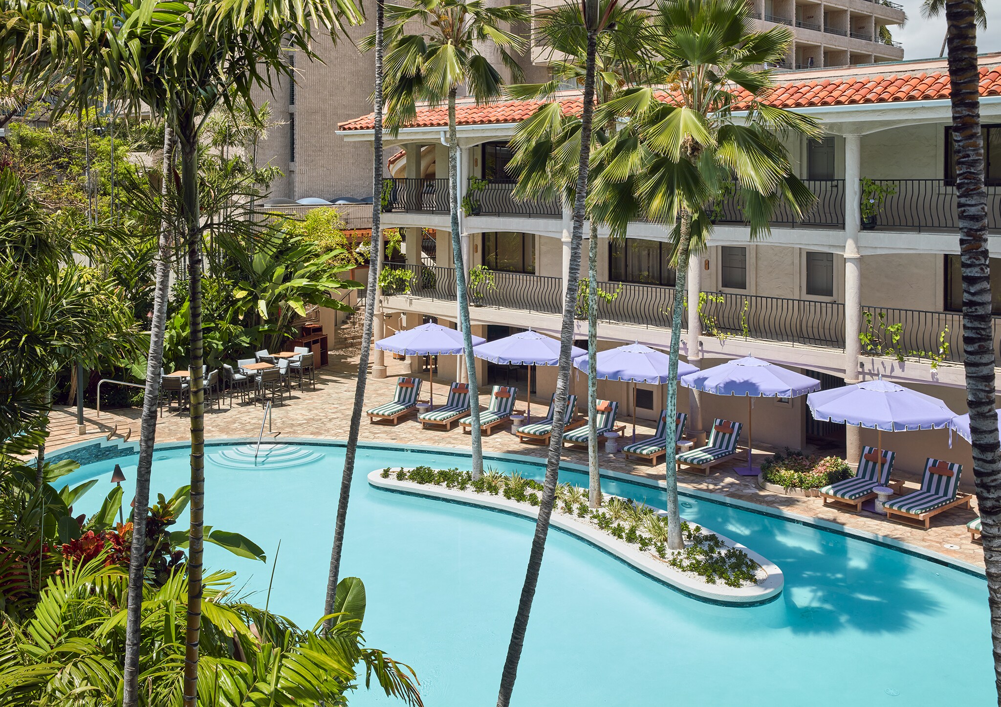 A saltwater lagoon pool is punctuated with a small island with three palm trees. Sun beds with purple umbrellas line the pool.