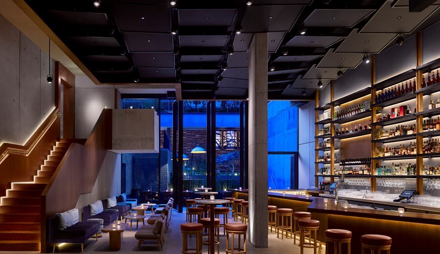 The Nobu Bar has high ceilings, a zigzag bar, stairs leading up one wall with embedded lighting, and a glass wall opening into a courtyard.