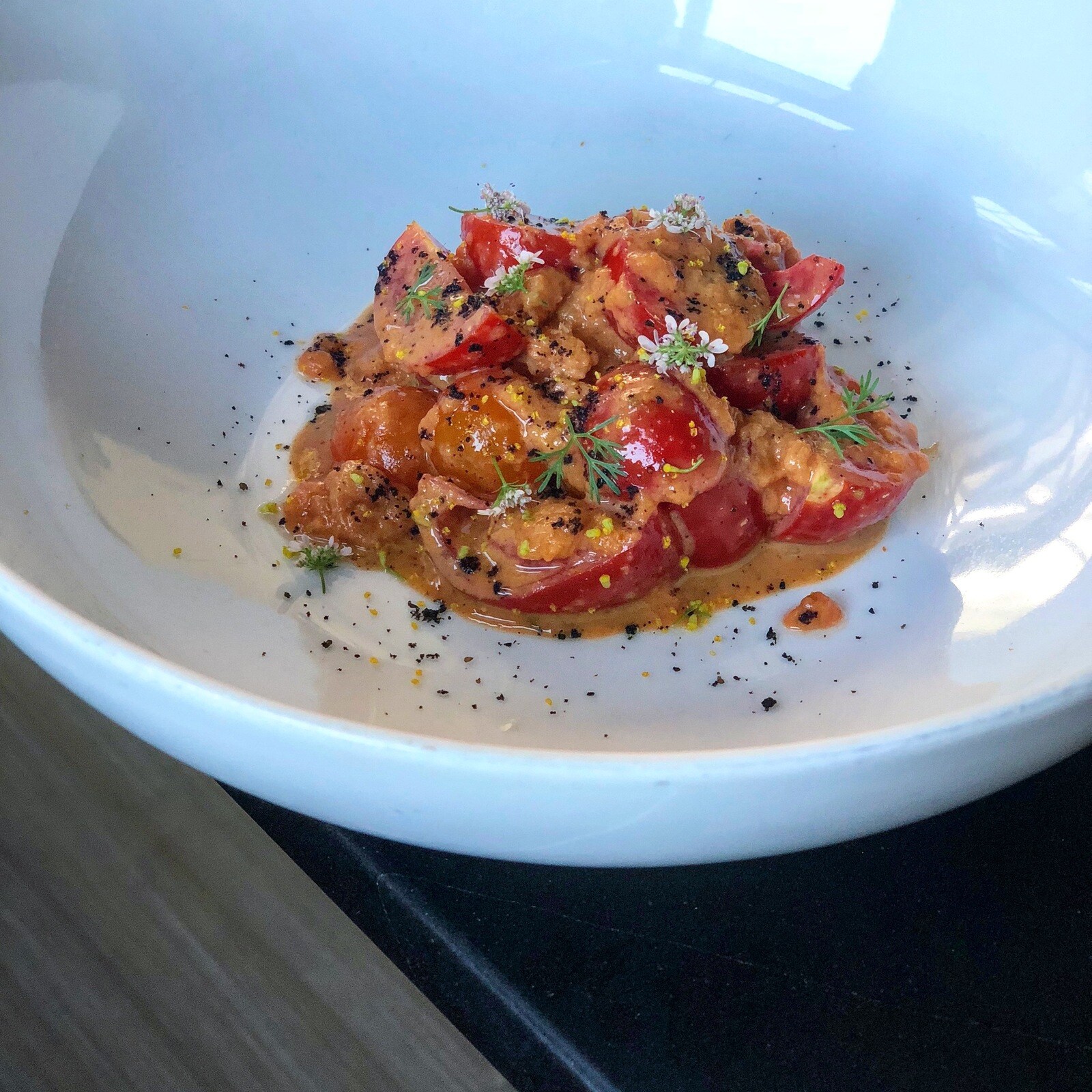 In a white bowl, a pile of tomato wedges sits in a light amber broth, topped with tomato chutney, sprigs of herbs, and tiny white flowers.