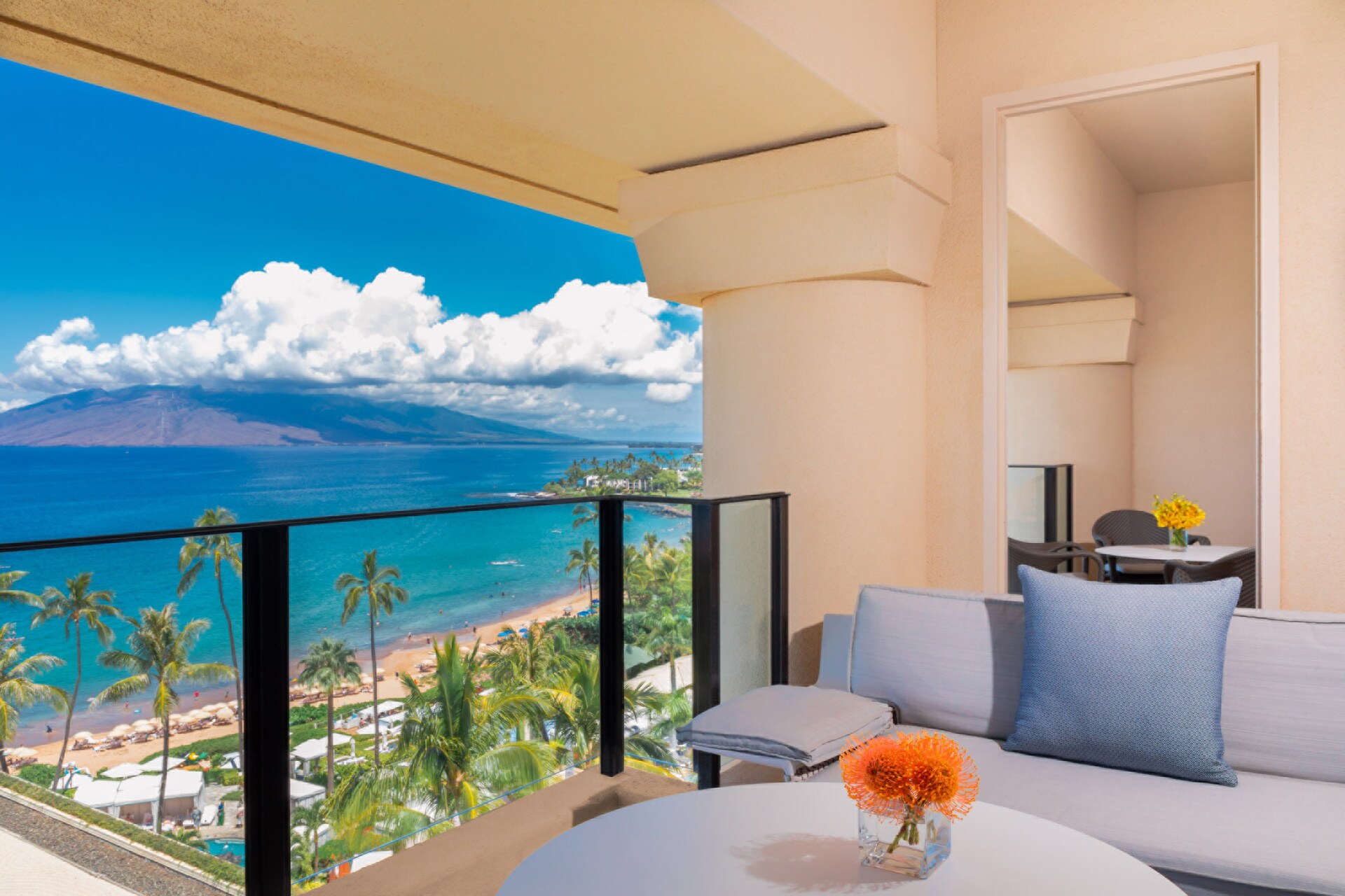 A blue couch sits next to a round table and on a balcony, which looks out on palm trees, a sunny strip of beach, and bright blue water.