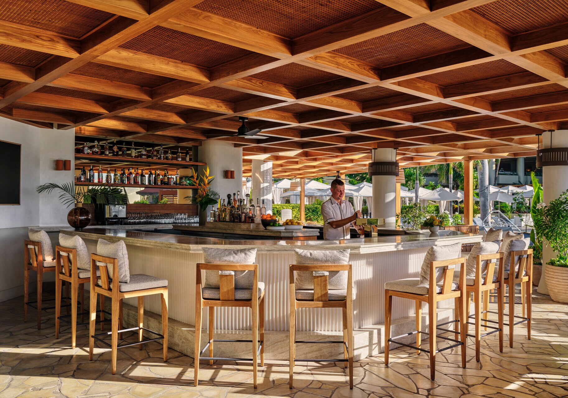 In a semi-outdoors bar with tan stone floors, high stools, and light gray counters, a white male bartender pours a drink from a tap.