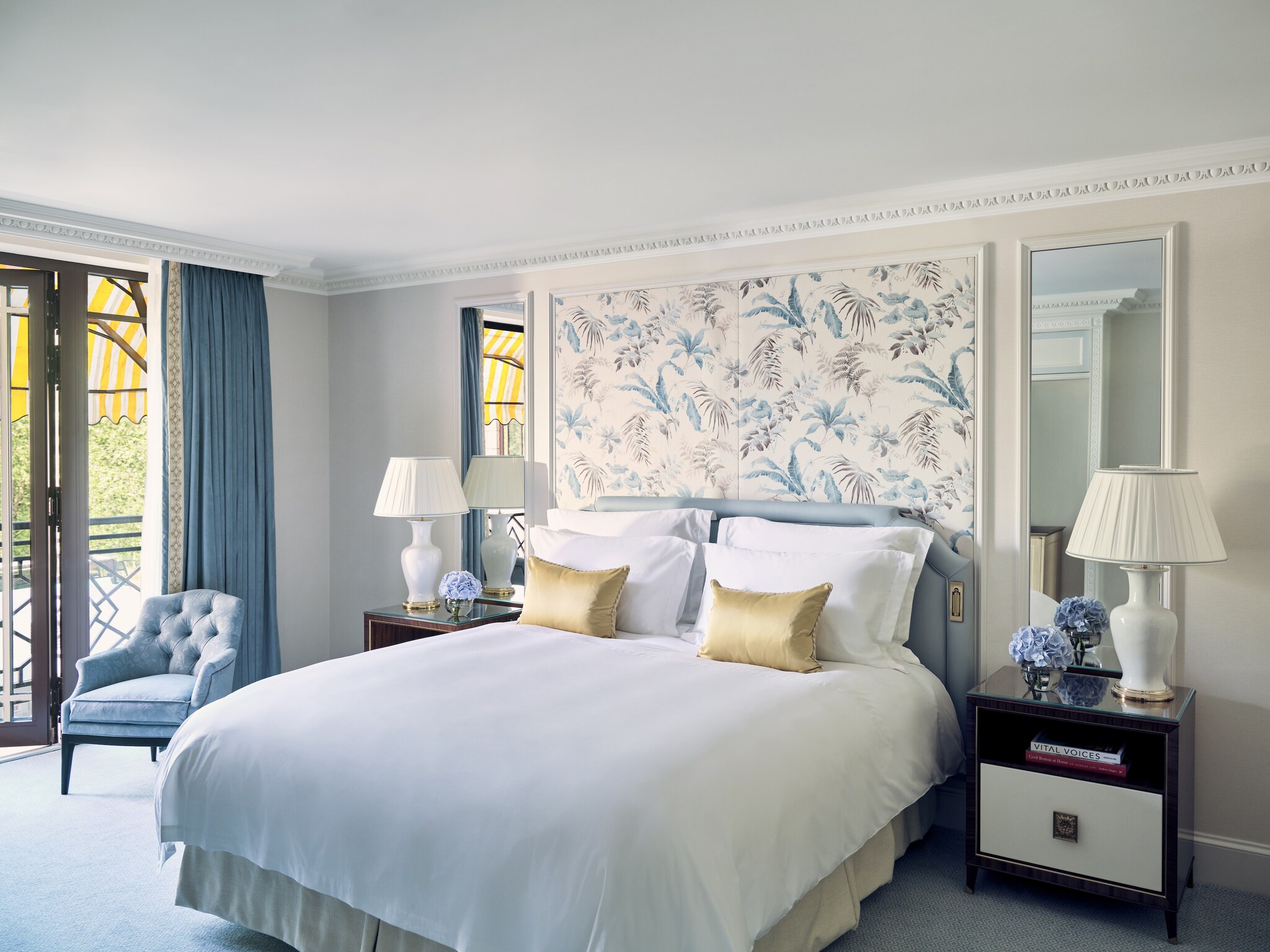  A hotel room is decorated in light blues, with a floral wallpaper backing the bed. Along one wall, glass windows open onto a private patio.