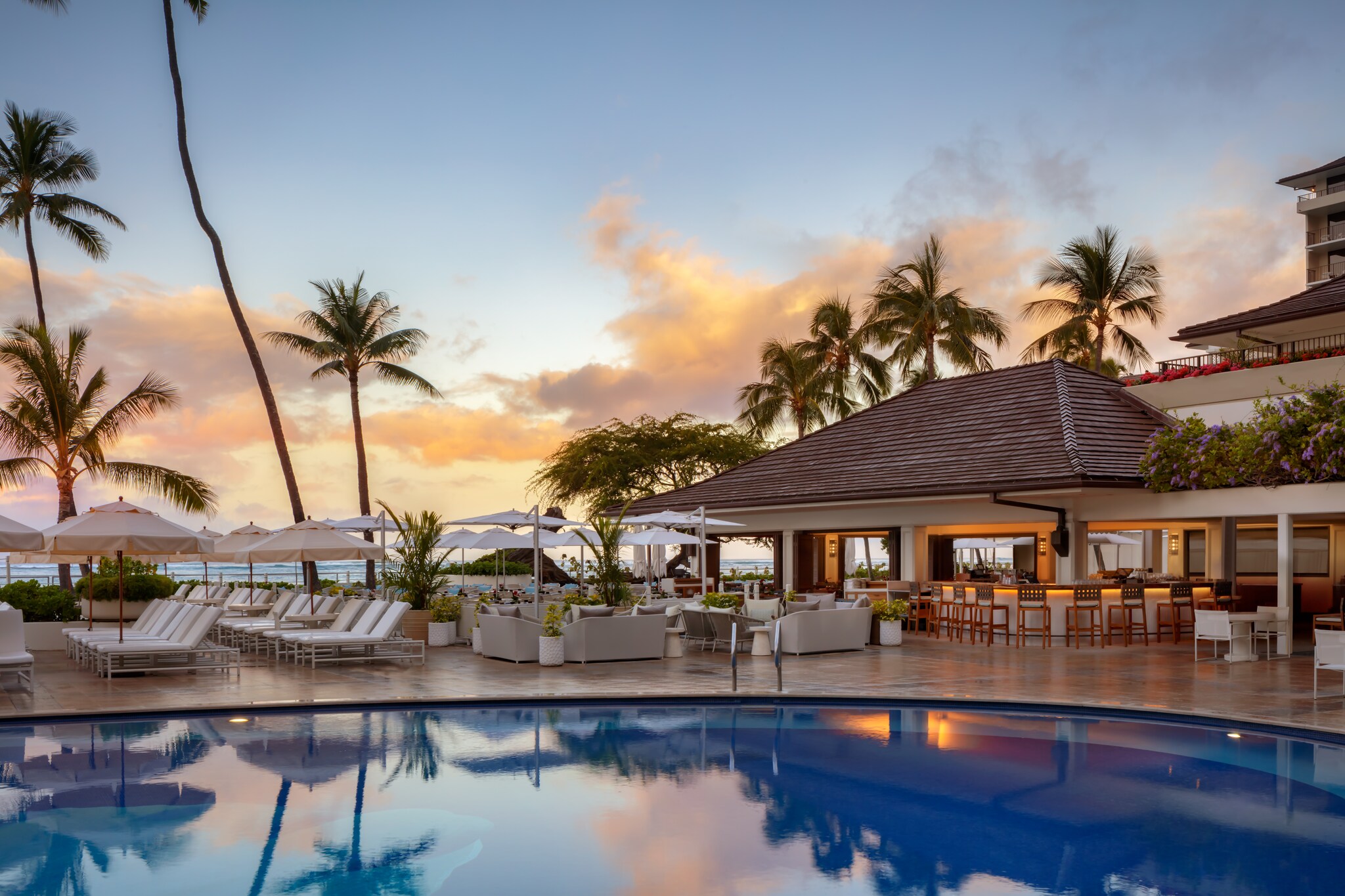 In the early evening, the hotel pool reflects the sky. Sun beds and umbrellas are arrayed in rows and the hotel is lit for the night.