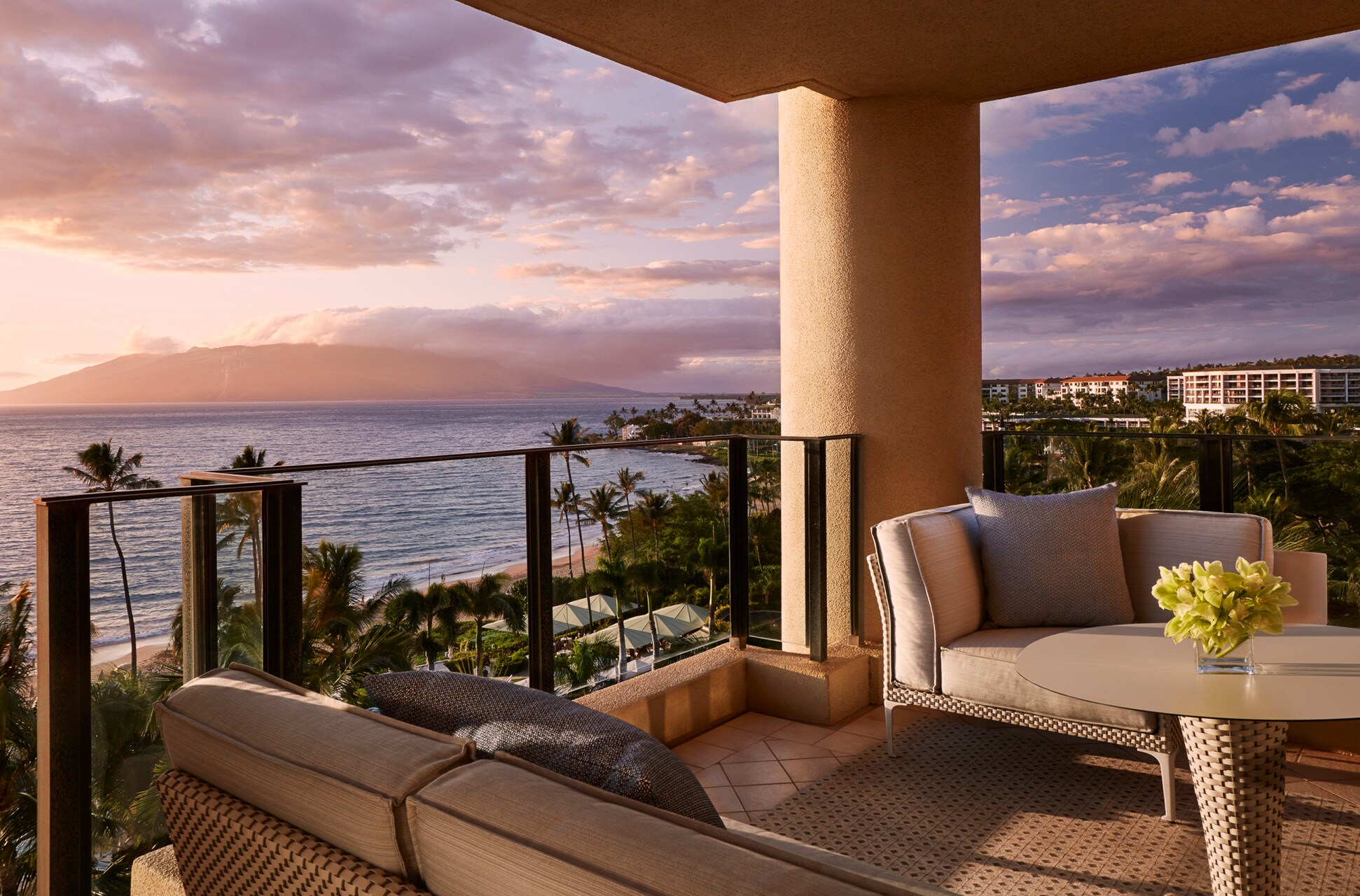 The balcony of the Ocean Front Prime suite has a small table, couch, and armchair. Beyond the railing, sunset shines pink over the beach.