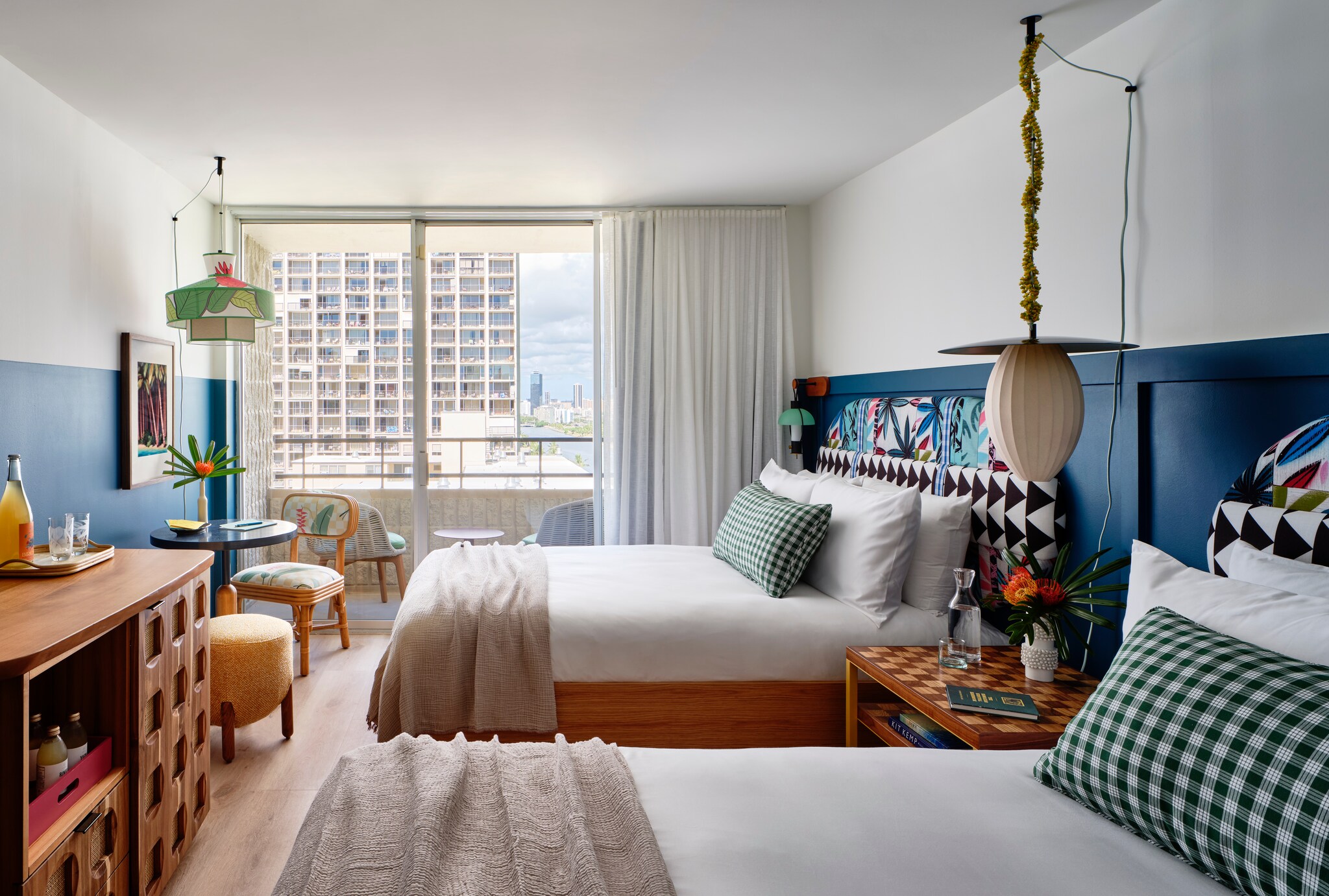 A room with two double beds is painted white and dark blue, with patterned headboards and floral accents. The far wall opens onto a lanai.